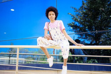 Full body of young African American female on overall looking at camera while sitting on railing near glass building in city