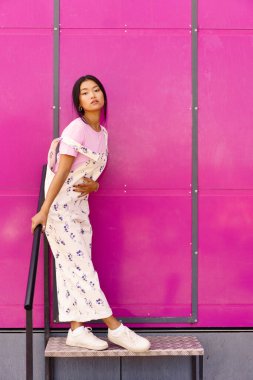Full body stylish Asian female in casual clothes touching belly and looking at camera while leaning on banister near bright pink wall