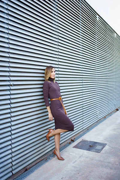 Full body female entrepreneur in stylish dress and high heeled shoes leaning on metal wall of modern building on city street