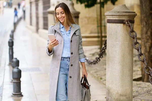 Cheerful adult female in stylish gray coat with bag smiling and browsing social media on cellphone while standing near pole with chains on city street
