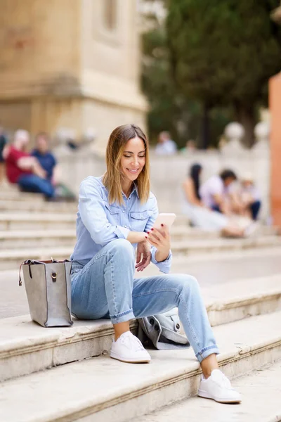 Full body positive adult female in casual clothes smiling and using mobile phone, while sitting near bag on stairs outside historic building in daytime in city