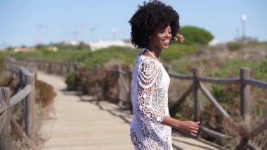Cuban woman with afro hair, dancing on a wooden path near the beach. Positive female with joy doing Cuban salsa moves.