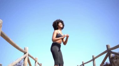 Positive black woman with afro hair jumping between wooden fences on a path to the beach, while playing sports on vacation. Slow motion video