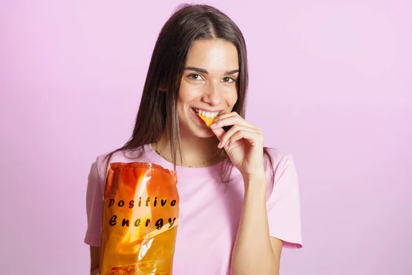 Positive young brunette in colorful t shirt standing on pink background and eating tasty crisps from orange packaging with Positive Energy inscription