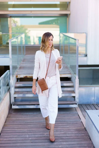 Full body woman in stylish clothes strolling on boardwalk and using mobile phone outside contemporary building