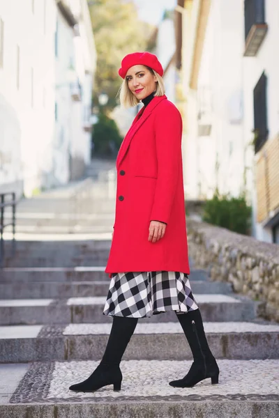 Full body of positive female in stylish red outfit looking away while walking on stone steps on streets with buildings
