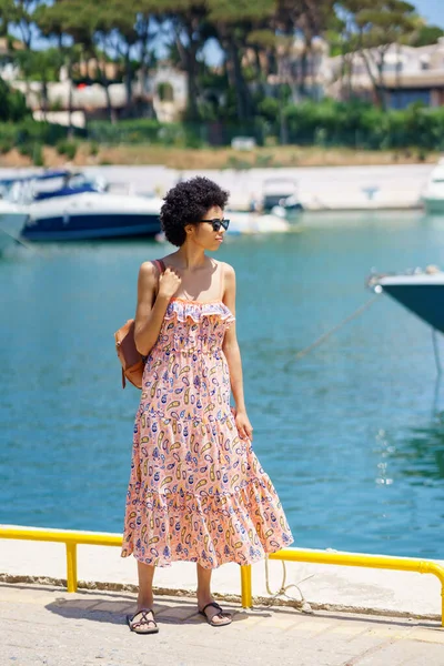 Full body black woman in long dress and sunglasses carrying bag on shoulder, and admiring sea while standing on pier near boats on summer weekend day in town port