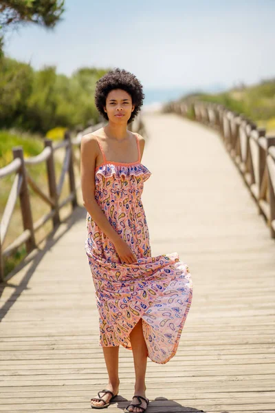 Full body young black woman with curly hair looking at camera, and waving skirt of long dress while standing on boardwalk leading to sea on sunny summer day