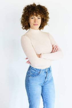 Self assured female model with dark curly hair in beige turtleneck and jeans crossing hands while standing against white background