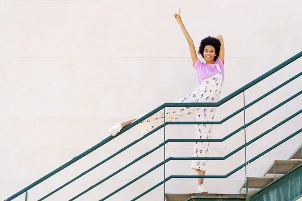 Full body of happy African American female, in casual outfit pointing up with raised arm and looking at camera while standing on stairway with leg on railing against white background
