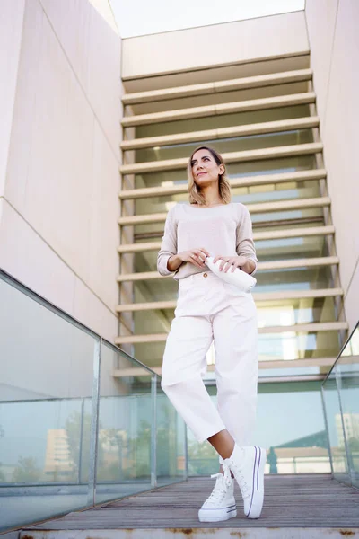 Low angle of woman in stylish outfit opening zero waste bottle of water and looking away while standing on path outside contemporary building