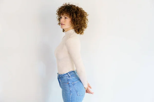 Side view of adult female in beige turtleneck and jeans standing with arms behind back against white background looking away
