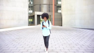 Positive young African American female student with afro hair, walking by her College faculty at the university, consulting her smartphone.