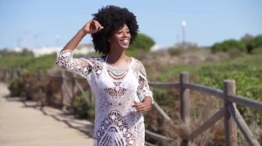Afro saçlı Afrikalı bir kadın, sahilin yakınındaki tahta bir patikada dans ediyor. Küba salsa hareketlerinden zevk alan pozitif siyah kadın..