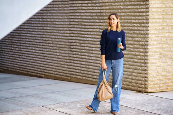 モダンな建物の外の舗装に立っている間に離れて見て袋と魔法瓶とスタイリッシュなジーンズとセーターで全身女性 — ストック写真