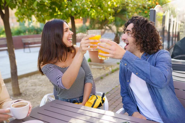 快乐的男女朋友 用橙汁碰杯庆祝夏天在咖啡店相遇 坐在桌旁互相望着对方 — 图库照片