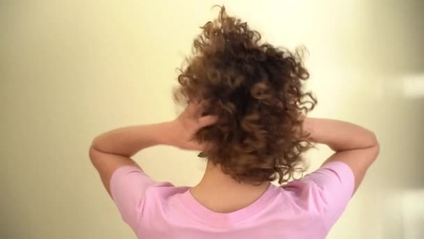 女性モデルのリアビュー 巻き毛と 彼女の頭の上に彼女の手で再生しながら カメラを見て ピンクのTシャツを着た白人女性 — ストック動画
