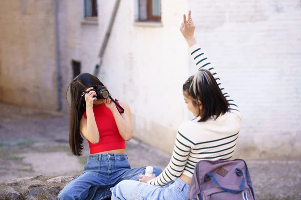 休暇中に街の路上で平和ジェスチャーを示すガールフレンドの写真を撮る若い女性写真家 — ストック写真