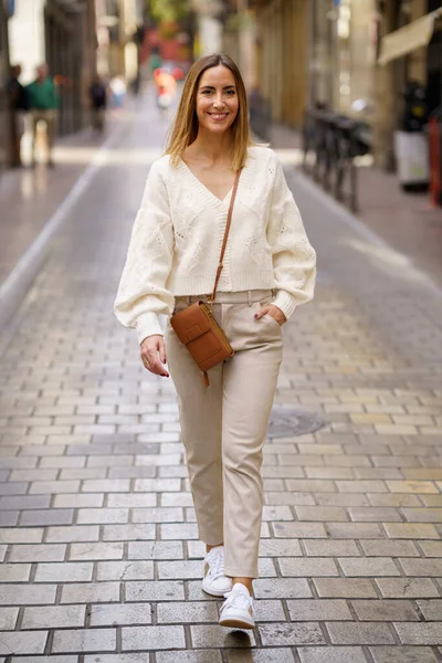 Full body of positive woman in beige clothes with brown shoulder bag walking on street with hand in pocket
