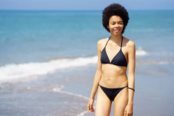 夏の日に熱帯リゾートの波に沿って海岸を散歩しながら距離を見て水着で陽気アフリカ系アメリカ人女性 — ストック写真