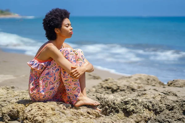 在暑假期间 穿着装饰玛西裙的赤脚黑人妇女坐在汹涌的岩石上 靠着汹涌的大海 抱着膝盖 望着远方 — 图库照片