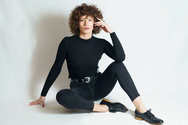 Full body of young female model, in trendy outfit relaxing with support of hand on floor and touching cheek with fingers while leaning on raised leg and looking at camera.