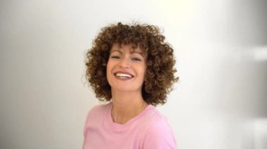 Kıvırcık saçlı çekici genç kadın model, elleri kafasında oynarken kameraya bakıyor. Pembe tişört giyen beyaz bir kadın.
