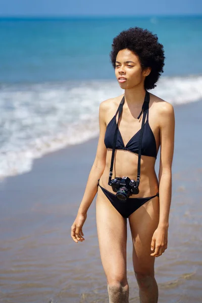 夏の日に海の近くの海岸を歩く写真カメラと水着でアフリカの髪型と深刻なアフリカ系アメリカ人女性写真家 — ストック写真
