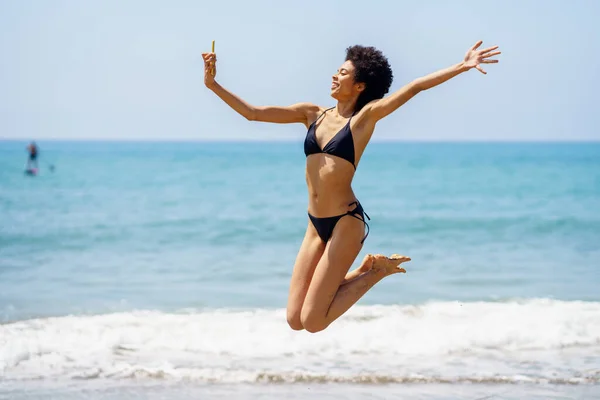 フルボディサイドビューの陽気アフリカ系アメリカ人女性旅行者でビキニ取ります自己肖像画上のスマートフォンと上の海岸でジャンプ近く手を振っ海 — ストック写真