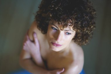 Kıvırcık saçlı yüksek açılı bir kadın, çıplak omuzlarıyla pencereden dışarı bakıyor ve kollarını kavuşturup yerde otururken bakışlarını kaybediyor.