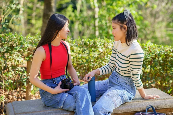 コンテンツ若いアジアの女性の友人 アルハンブラの森の中でハイキング旅行中にルートを議論しながら カメラと魔法瓶とベンチに座ってカジュアルウェアで — ストック写真