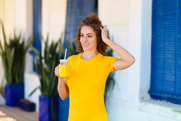 幸せ若いです女性で黄色Tシャツで巻き茶色の髪笑顔と離れて見て 新鮮なジュースのガラス瓶で通りに頭を触れながら — ストック写真