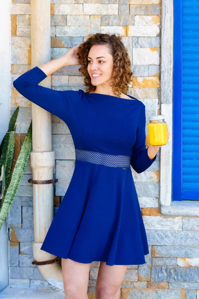 ポジティブな若い女性で青いドレスに触れ巻き毛と離れて見ながら立っている間にガラス瓶のオレンジジュースでレンガ造りの建物に対して — ストック写真