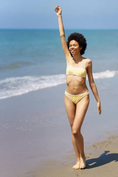 明るい若いアフリカ系アメリカ人女性のフルボディ 流行のビキニで暗い巻き毛を持つ旅行者 笑顔と太陽の下で腕を上げ砂のビーチを歩きながら離れて見て — ストック写真