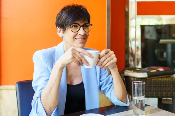 流行の青いジャケットと眼鏡の正の女性は熱い飲み物のカップとテーブルに座って カフェテリアで離れて見て — ストック写真