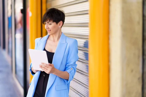 正式な服の正の女性起業家は 昼間のタブレットを閲覧しながら 通りに黄色の列と銀の圧延ドアで立って — ストック写真