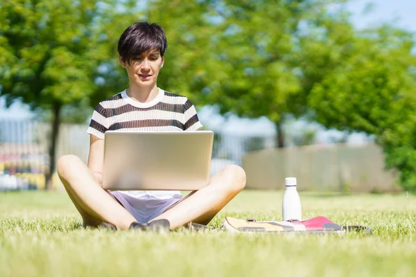 草の上に座って 公園でノートパソコンでリモートで作業し 宿題をしている間に画面を見てカジュアルな服の女性のフリーランサーの完全な体 — ストック写真