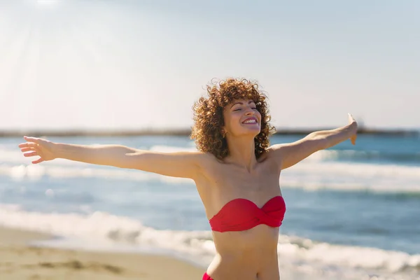 穿着粉色比基尼的快乐的年轻红头发女性 双目紧闭 张开双臂笑着 站在沙滩上 面对蓝色的海水和天空 阳光普照 — 图库照片