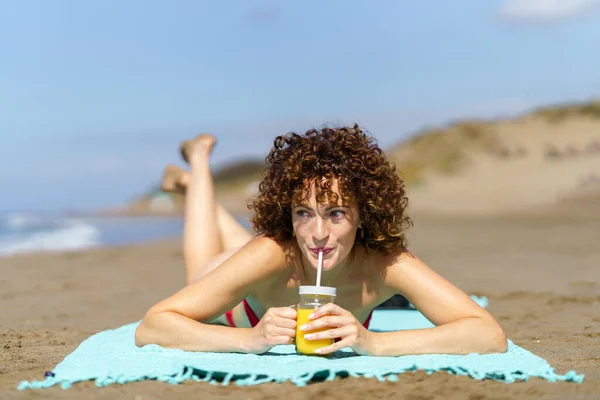 年轻的女性 一头卷曲的红头发躺在海滩上 一边喝着杯子里的橙汁 一边望着旁边的稻草 — 图库照片