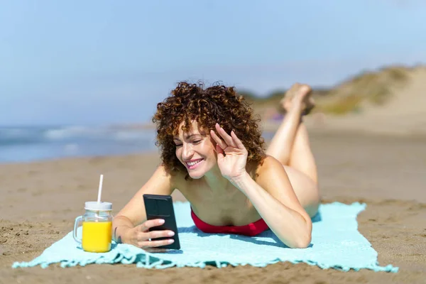 年轻快乐的女性 长着姜黄卷发 用橙汁躺在沙滩上 一边挥手一边打电话一边视频 — 图库照片