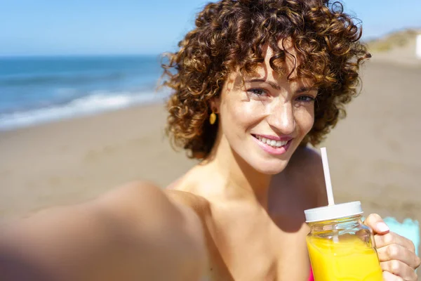 夏休みにビーチで自撮りしながら爽やかなオレンジジュースを飲む巻き毛の若い女性の笑顔の肖像 — ストック写真