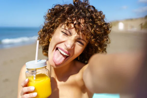带着姜黄卷发的爱开玩笑的女人 一边拿着一杯橙汁 一边在海滨自拍 一边伸出舌头 — 图库照片