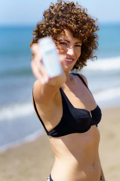 身穿比基尼的年轻女性微笑着站在海浪边的沙滩上 展示着一瓶白色的防晒霜 — 图库照片