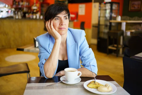 喫茶店で腕を傾けている間に飲み物やケーキのカップとテーブルに座ってエレガントなジャケットで思慮深い女性 — ストック写真