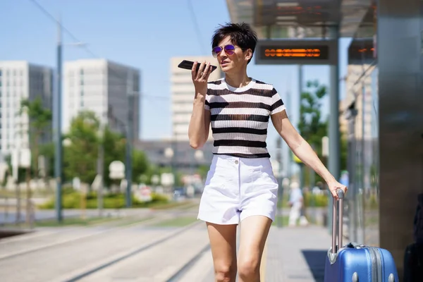 流行の衣装を着た前向きな若い女性と 鉄道で街の通りに立つスーツケースを着たサングラス そして明るい日差しの中で携帯電話にオーディオメッセージを記録する — ストック写真