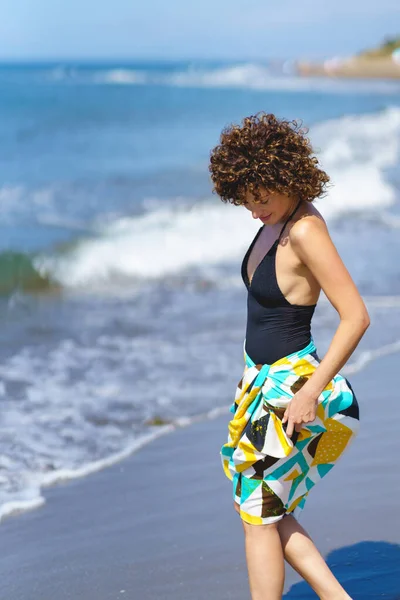 カラフルなラッパー布とカジュアルな上に巻き毛の若い女性の側のビューは 立って 日光で海を振って近くの砂浜を見下ろします — ストック写真