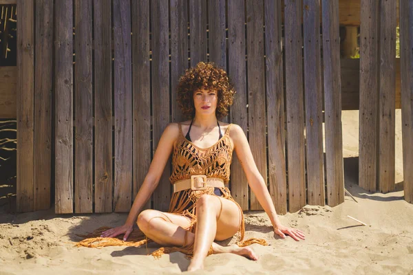 若いです裸足の女性の完全な体とともに巻き毛で茶色のかぎ針編みのビーチカバーアップ座って上の砂の上に傾いて晴れた日に木製のフェンス — ストック写真