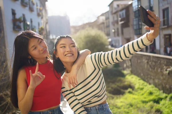 幸せなアジアの女性ショーホーンジェスチャーとととととともに手の肩の友人の上に自分の肖像画を取りながらスマートフォンで一緒にグラナダ — ストック写真