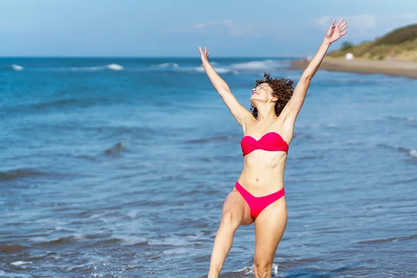 穿着粉色比基尼的快乐的年轻女性 一边享受暑假 一边挥动着海水 举起双臂 — 图库照片