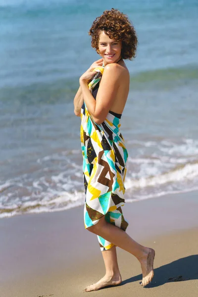 正の若い女性のフルボディ側のビューでカラフルなドレスは日光で海を振って近くの砂浜に立って カメラを見て — ストック写真